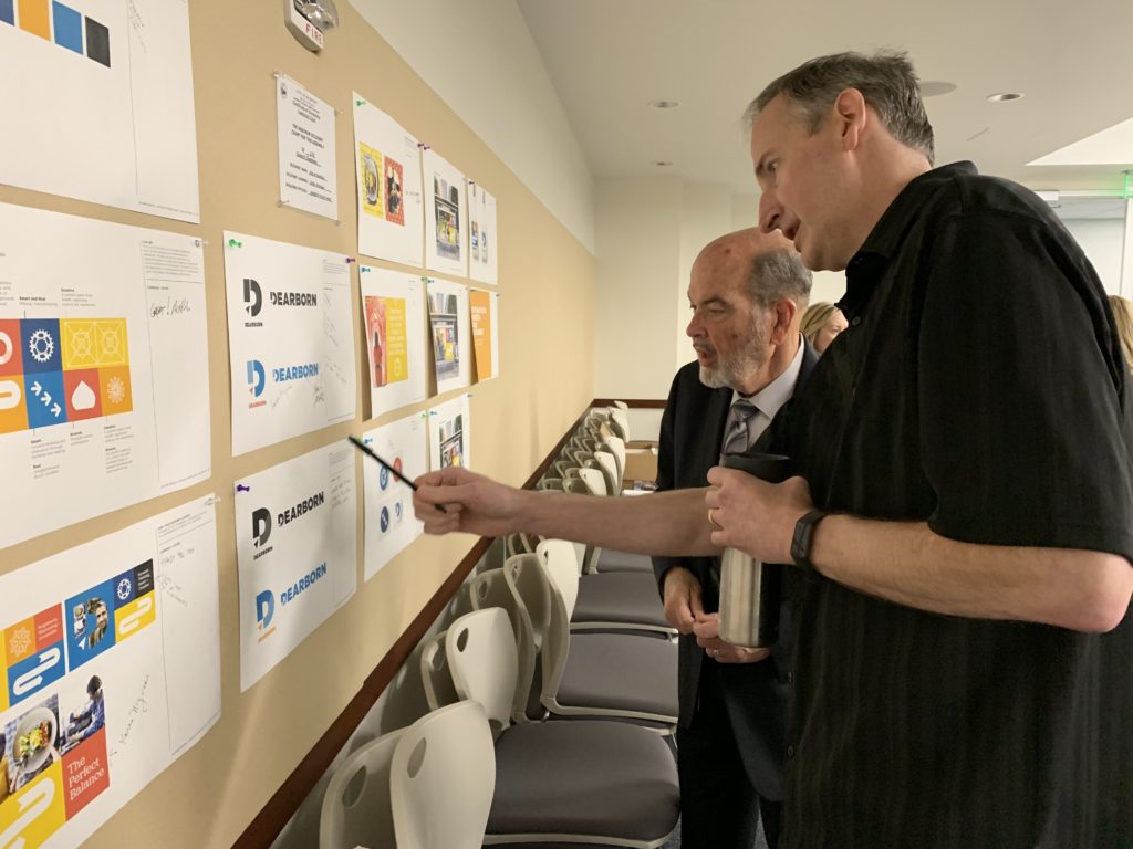 Two men reviewing printed concept proofs of Dearborn branding on a corkboard
